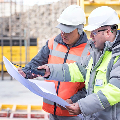 Two men in construction vests and hard hats looking at a paper.